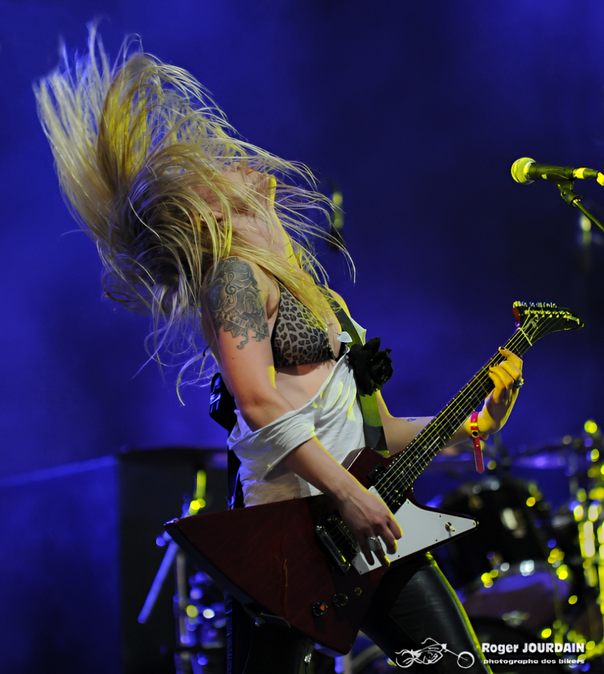 Gros plan sur une guitariste blonde sur scène en pleine performance, agitant ses cheveux (headbanging) lors d'un concert au Free Wheels.