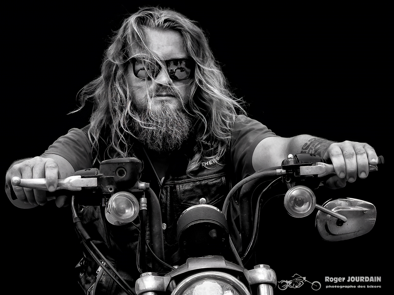 Portrait en gros plan d'un motard barbu aux longs cheveux et lunettes de soleil, sur sa moto