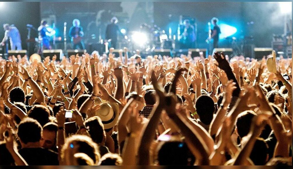 Foule enthousiaste levant les mains devant une scène de concert en soirée. Ambiance rock.
