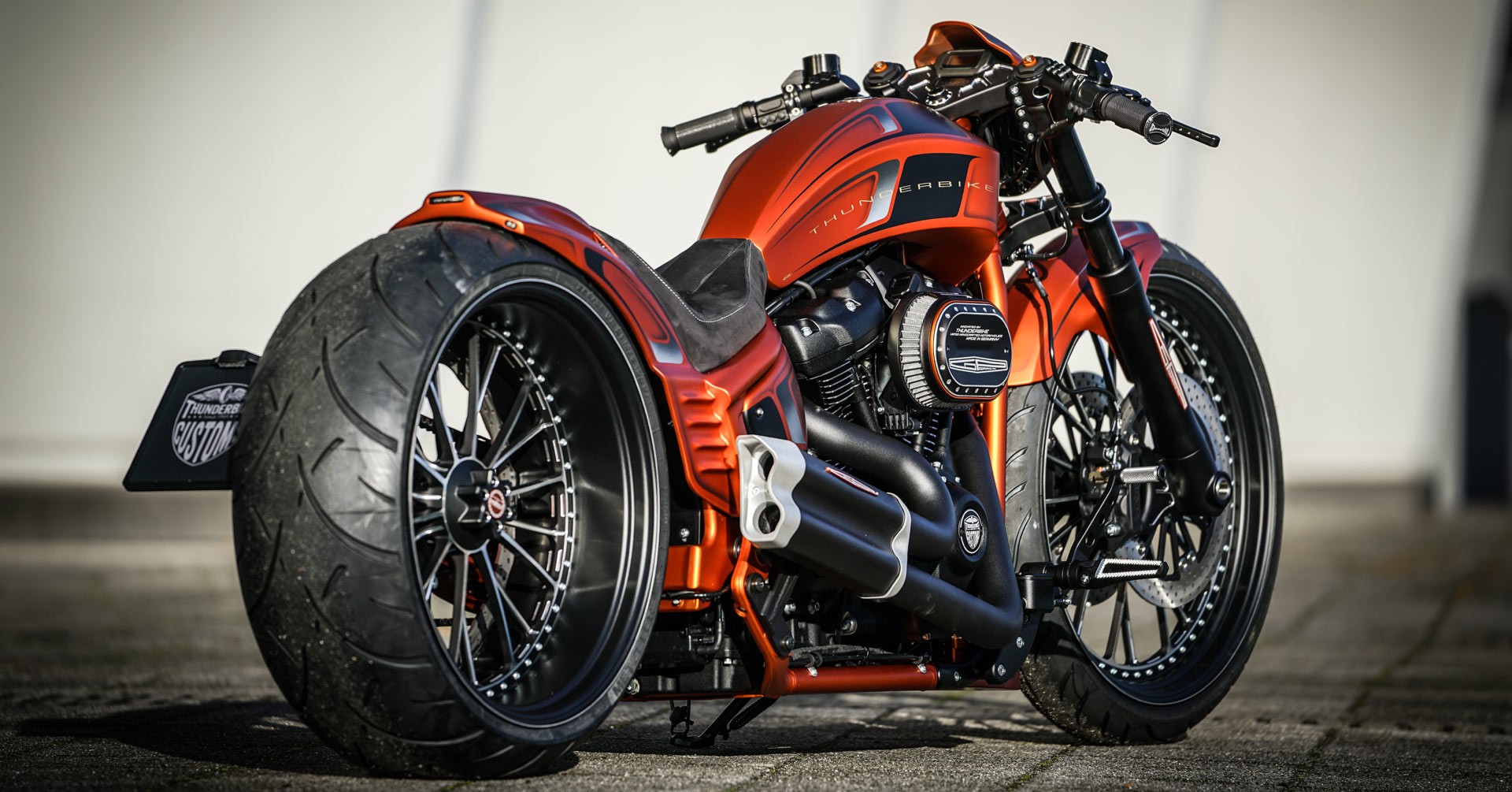 Moto customisée orange (chopper ou cruiser) avec gros pneu arrière, chromes et design agressif.